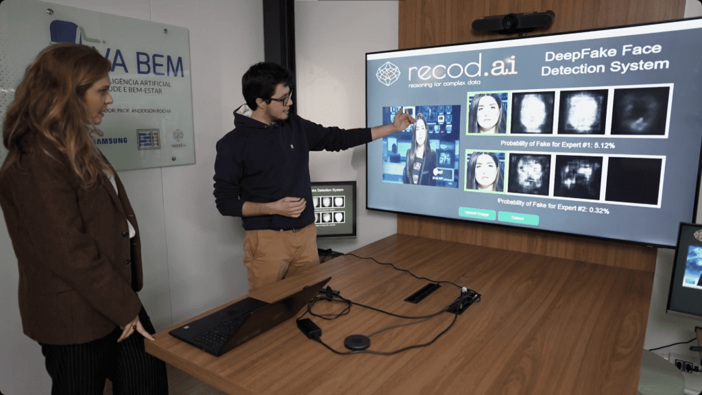 Pós-doutorando do Recod.ai, Gabriel Bertocco, apresentando à repórter do Fantástico a ferramenta que utilizam para detectar vídeos falsos gerados por IA. Ambos estão de frente pra uma televisão em que Gabriel apresenta a tecnologia.