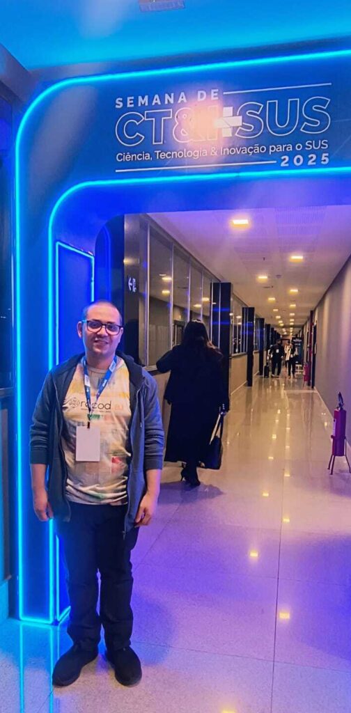 A imagem mostra Leopoldo Lusquino Filho, homem branco calvo em frente à entrada do evento "Semana de Ciência, tecnologia & Inovação para o SUS 2025" ocorrido em Brasília