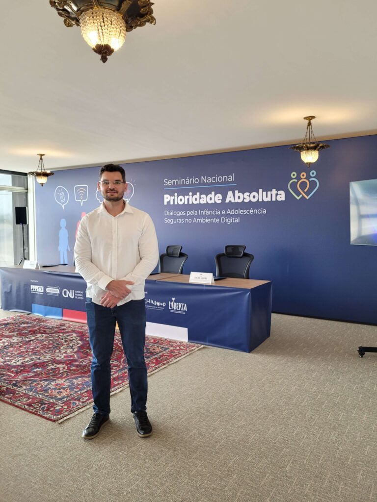 A imagem mostra o pesquisador Raphael Prates na frente de um painel com o nome do evento "Prioridade Abosulta". Prates é homem alto de óculos, cabelos castanhos escuros, de óculos. Ele está usando camisa branca e calça jeans.