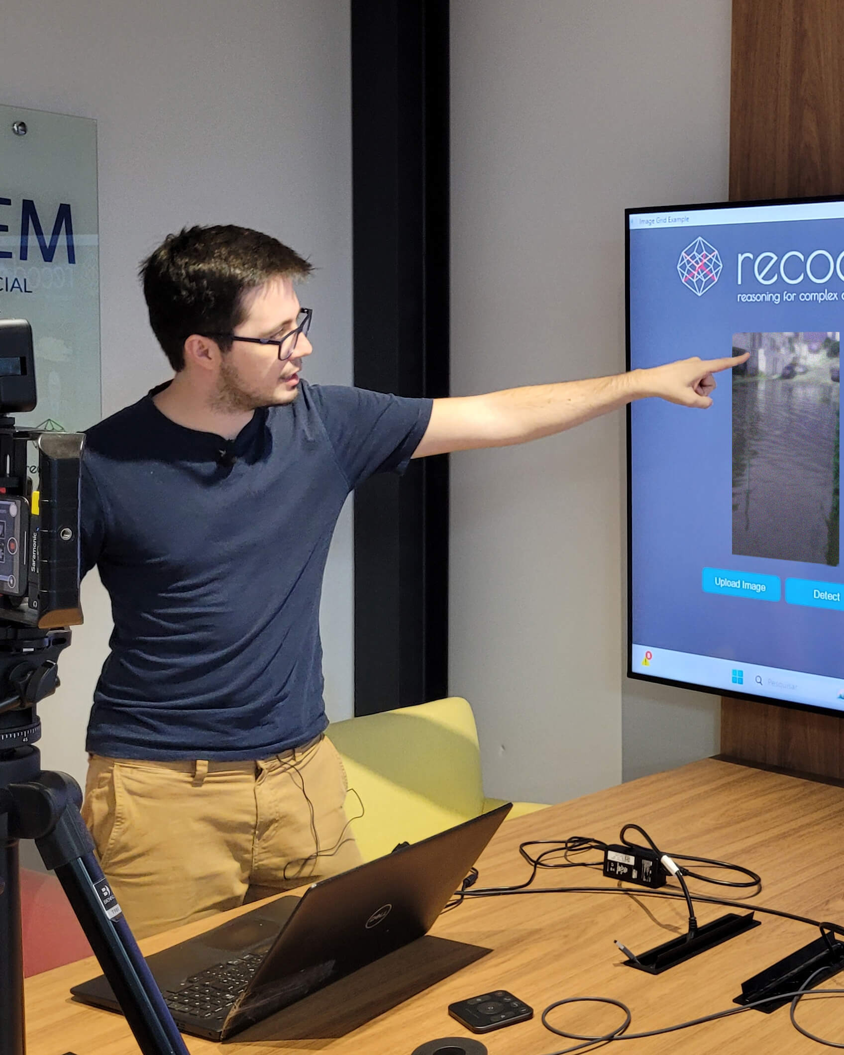 Gabriel Bertocco, homem branco, cabelos castanhos escuros curtos, de óculos, camiseta azul e calça marrom, apontando para uma imagem de enchente em um televisor.